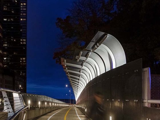 A curved pedestrian and bike path at night is illuminated by modern lighting, with a wave-like security screen and city buildings in view.