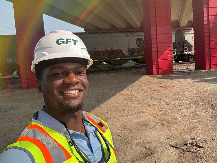 A GFT intern wearing a hard hat and high-visibility safety vest stands beneath an elevated rail structure near train tracks during a site visit.