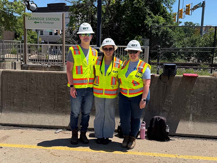 Three GFT interns wearing hard hats and high-visibility safety vests stand near a transit station platform during a field visit.